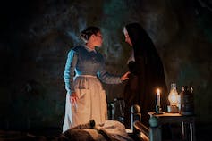في غرفة مضاءة بالشموع المظلمة ، تضع فلورنس بو (في ثوب أزرق ومئزر أبيض) يدها على ذراع راهبة.